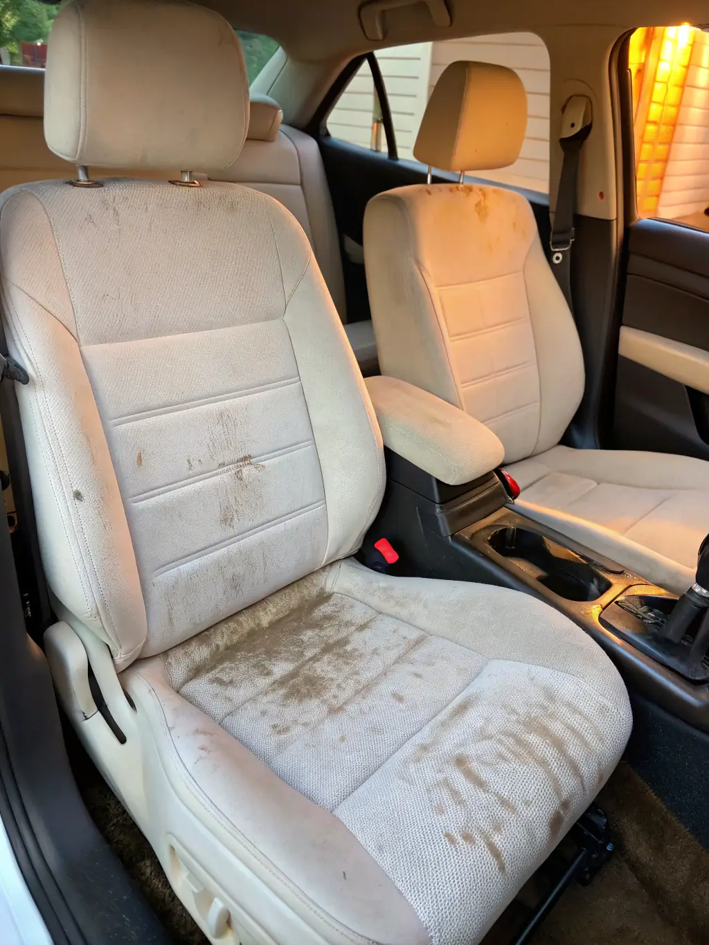 A professional photo of a bottle of 'Fabric Cleaner' with a brush applicator, being used on a car seat. The seat is visibly stained on one side and clean on the other, demonstrating the product's cleaning power.